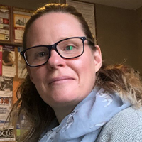 Community and Justice Services graduate Linzi Caldwell with brown hair tied back, wearing glasses and a blue scarf