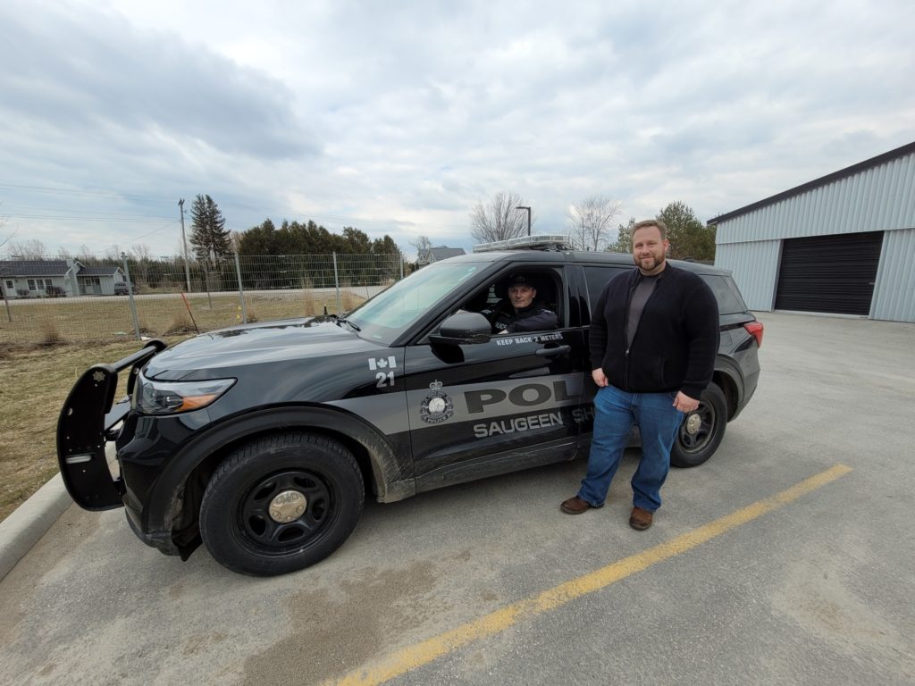 A white male with brown hair and a beard standing beside a police cruiser with a male police officer inside