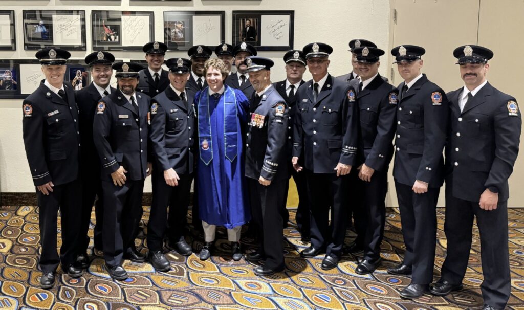 A large group of firefighters in uniform pose with a Georgian graduate in blue gown.