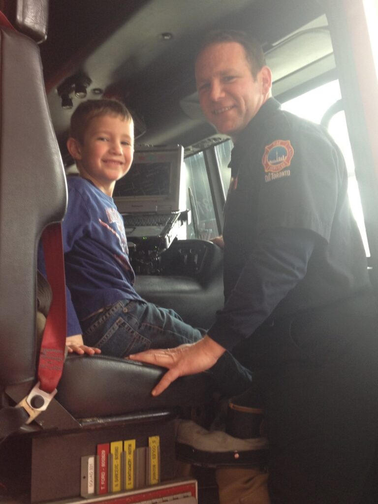 A child sits on a seat inside a fire truck with an adult standing next to them. 