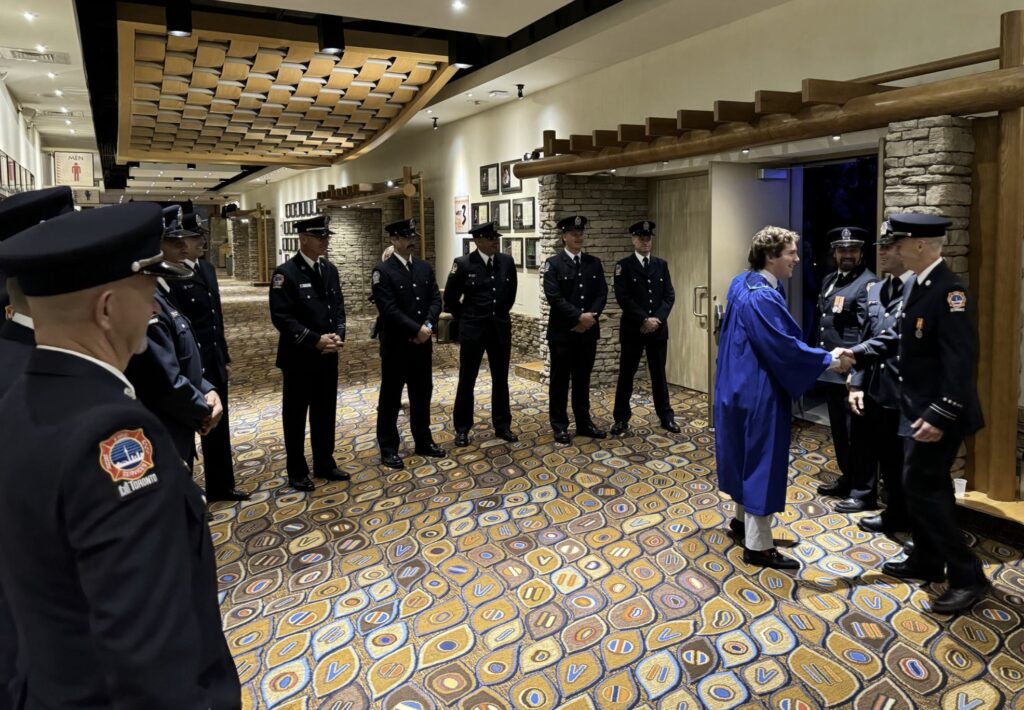 A semi-circle of firefighters in formal uniforms wait to shake hands with a person dressed in a blue convocation robe. 