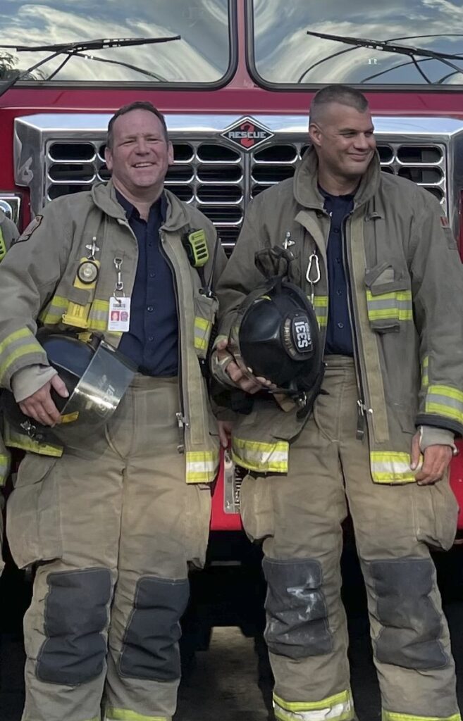 Two firefighters in uniform stand next to each other in front of a fire truck. 