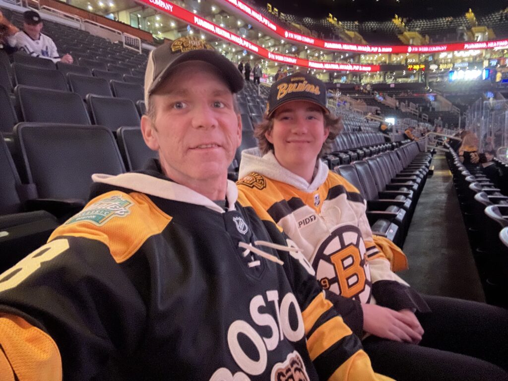 Two people wearing Boston Bruins sweaters and hats take a selfie inside a hockey arena.