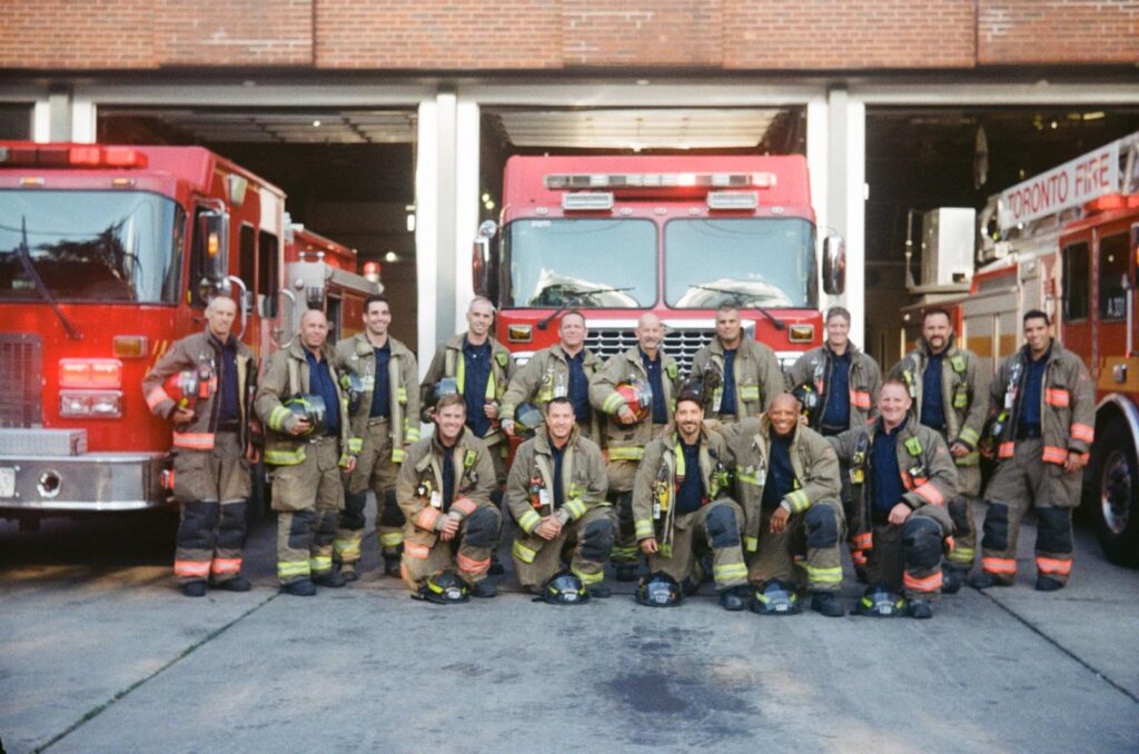 A group photo of firefighters in uniform outside of a fire hall with three fire trucks in the background. 