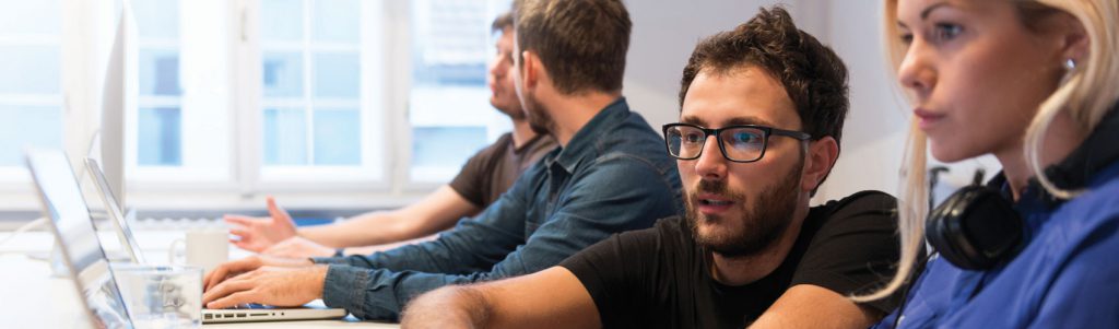A group of four Computer Programming and Analysis students sitting at a desk working on laptops in a classroom setting