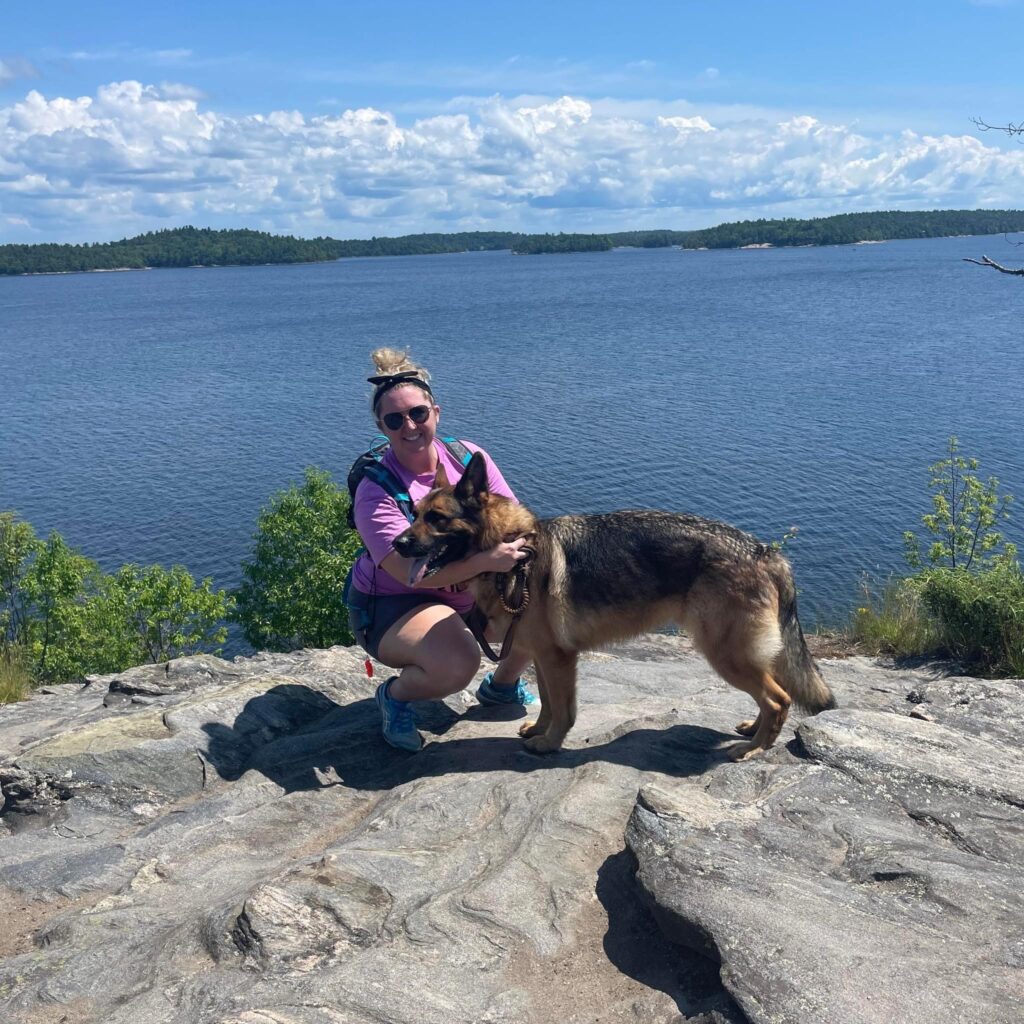 A person crouches next to a dog on a rock cliff overlooking a lake.