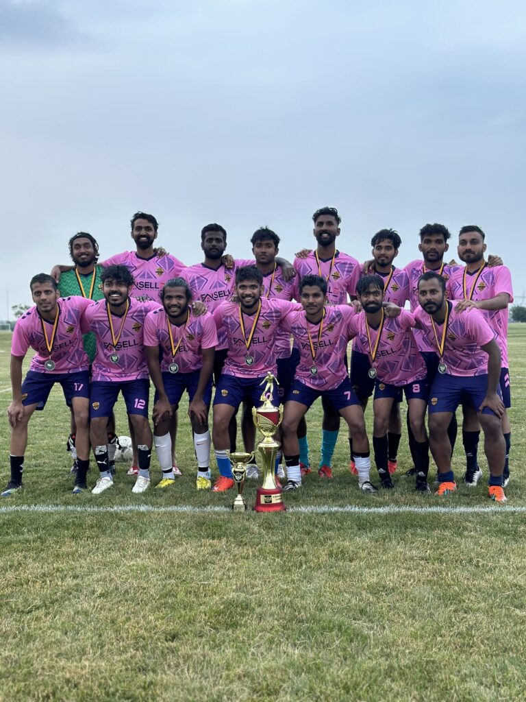 A group of people, wearing matching pink and blue soccer uniforms, pose on a soccer field with two trophies.