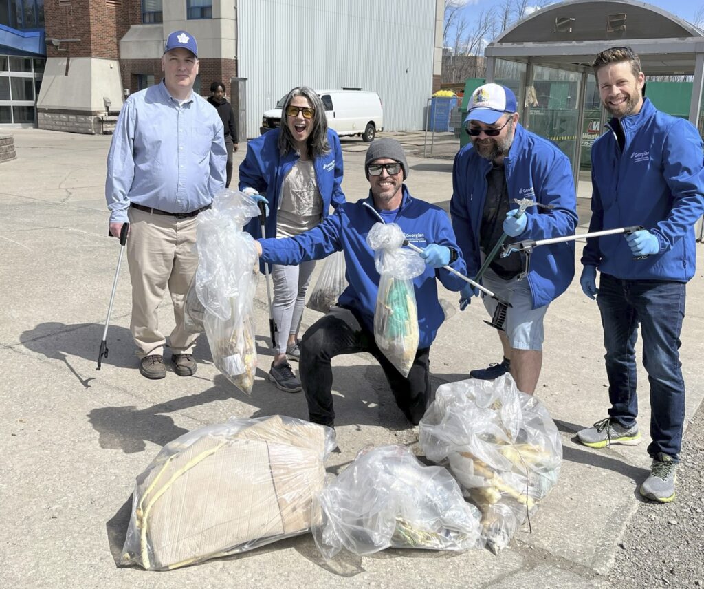 Five people wearing blue jackets and holding trash picker sticks smile and pose in front of a few bags of garbage. 