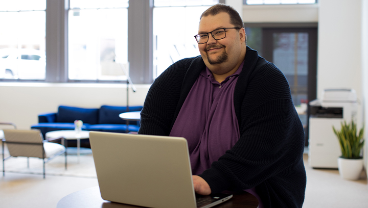 A person wearing black glasses, a purple golf shirt and black knit cardigan, sitting at a round table using a laptop in an office setting