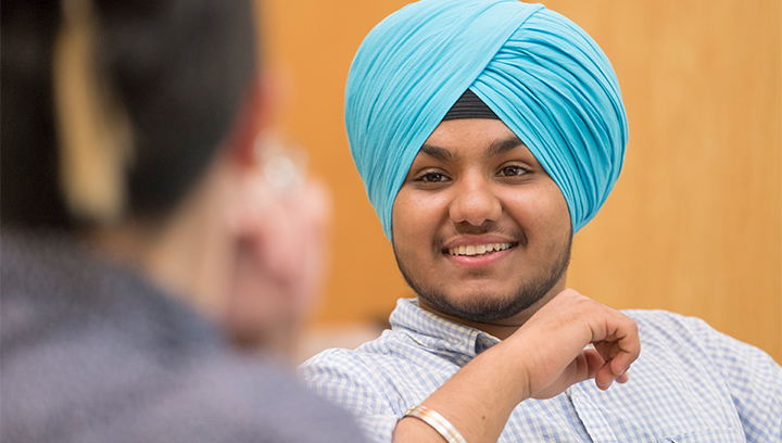 A person smiling, wearing a bright turquoise turban, white and light blue plaid shirt, and silver and gold bangle in conversation with another person