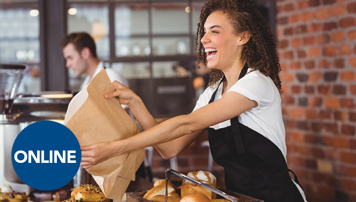 A food service worker with brown curly hair, in a white short-sleeved t-shirt and a black apron, smiling and holding out a paper bag in a cafe/bakery setting