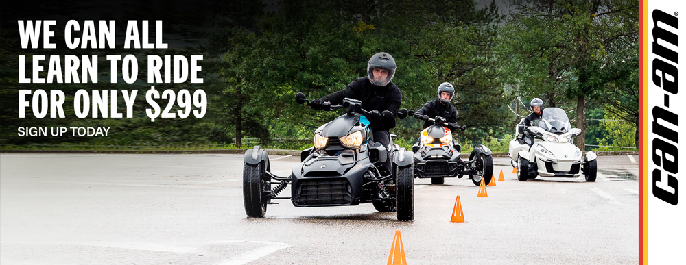 Two people wearing black jackets and helmets riding Can-Am three-wheeled motorcycles on asphalt around pilons with a forest in the background