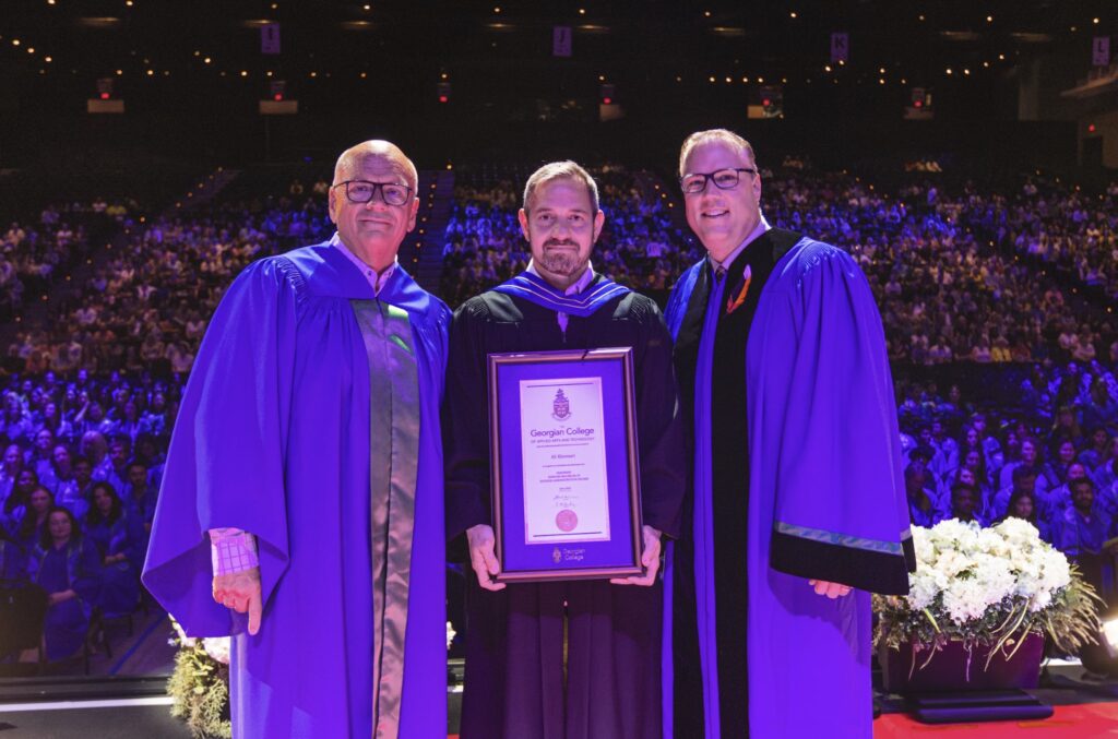 Three people wearing blue convocation robes smile and the person in the centre holds a framed certificate. They're standing on a stage, with a large crowd behind them.
