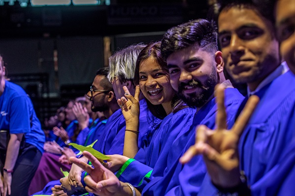 Graduates in blue gowns in the crowd smiling at the camera and holding up peace signs with their hands