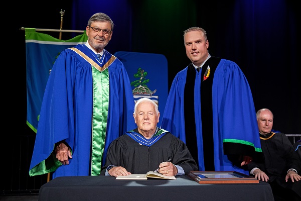 Peter B. Moore receives honourary degree; on stage with Georgian Board Chair and President - both standing over Peter as he signs a document