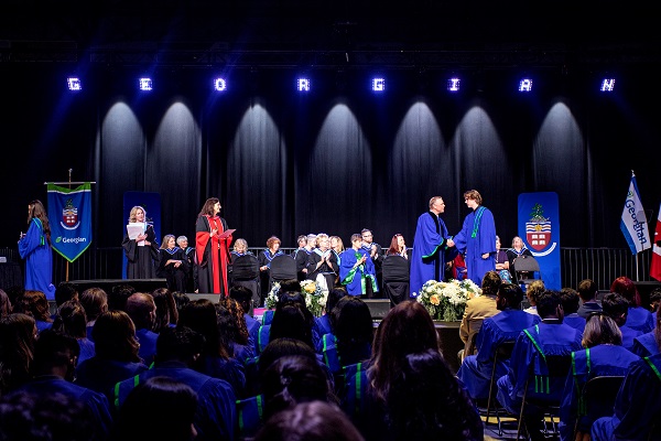 Wide view of stage at convocation, president shaking hand of graduate, both wearing Georgian blue gowns