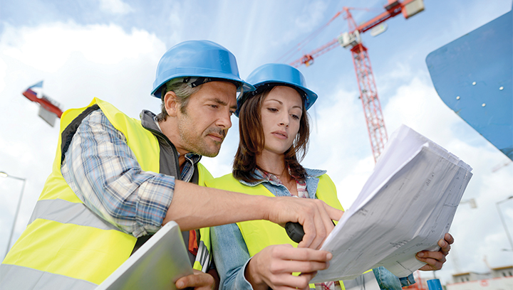 Two construction workers wearing bright yellow safety vests and blue hard hats, while looking at blue prints in front of a crane