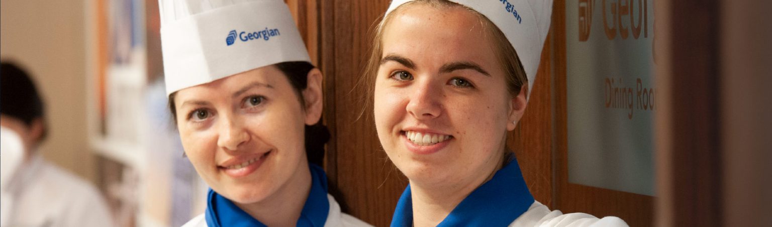 Two Culinary Management students wearing tall chef hats, uniforms and blue neck scarves in front of the Georgian Dining Room