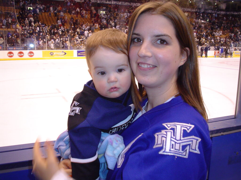 An adult holds a baby in a hockey rink. Both are wearing Toronto Maple Leaf jerseys. 