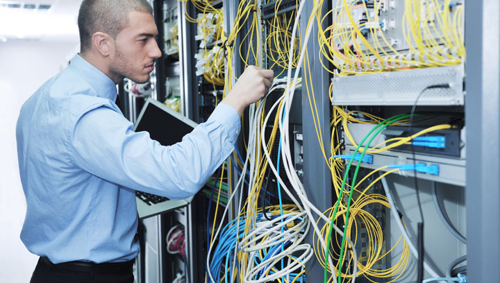 Cybersecurity specialist in a blue collared shirt holding a laptop and connecting wires to server equipment
