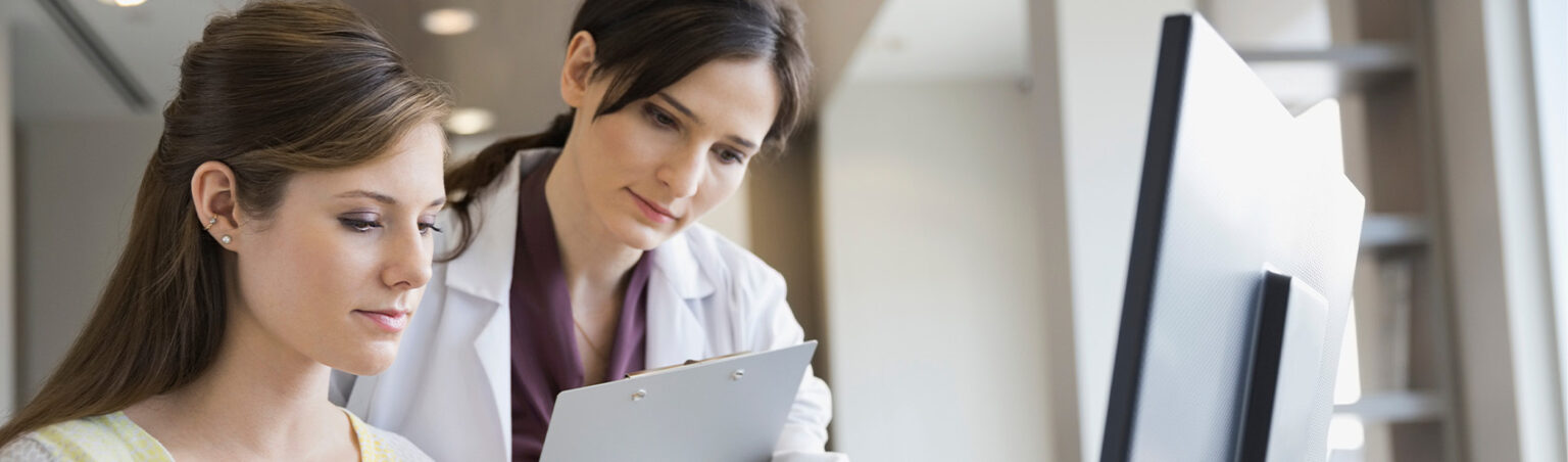 A healthcare professional holding a clipboard reviews information with a young woman seated at a computer.