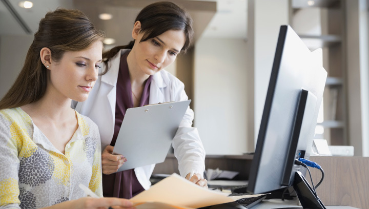 A healthcare professional holding a clipboard reviews information with a young woman seated at a computer.