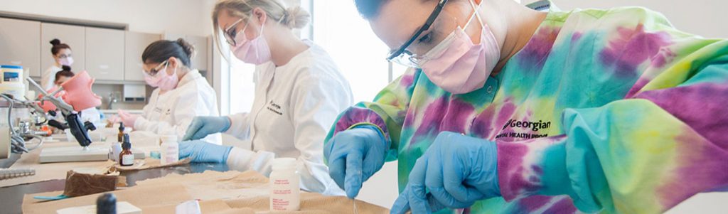Dental Assisting students wearing scrubs, safety glasses, masks and latex gloves preparing molds for patient mouth simulators