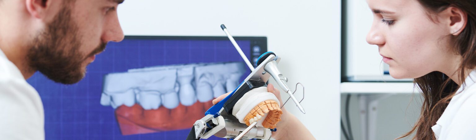 Two dental professionals examine a dental model with specialized equipment, with a digital image of teeth displayed on a computer screen in the background.