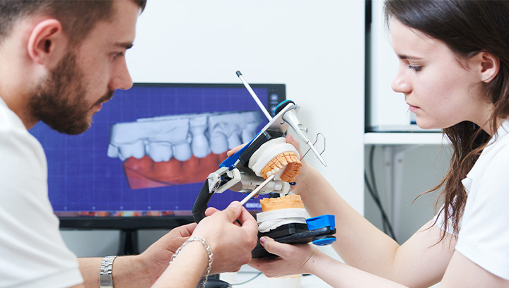 Two dental professionals examine a dental model with specialized equipment, with a digital image of teeth displayed on a computer screen in the background.