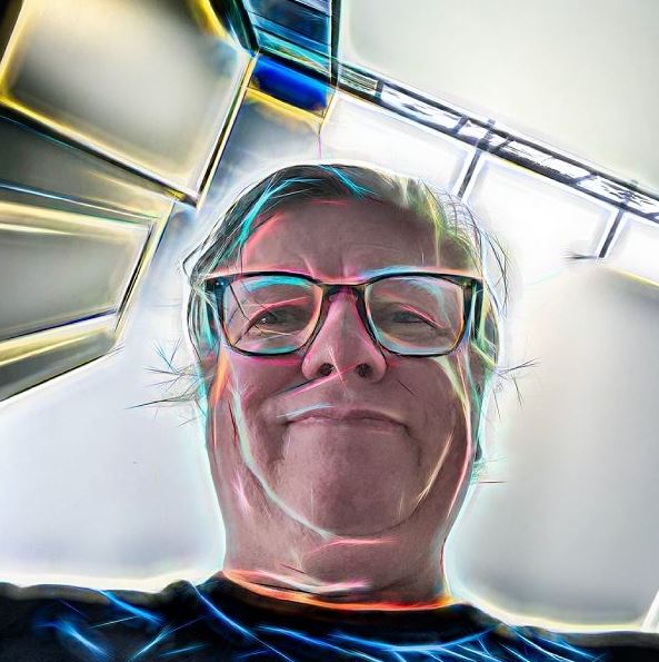 A selfie taken from below of a person with short hair, glasses and a dark shirt. The image has a variety of neon colours outlining different parts of it.