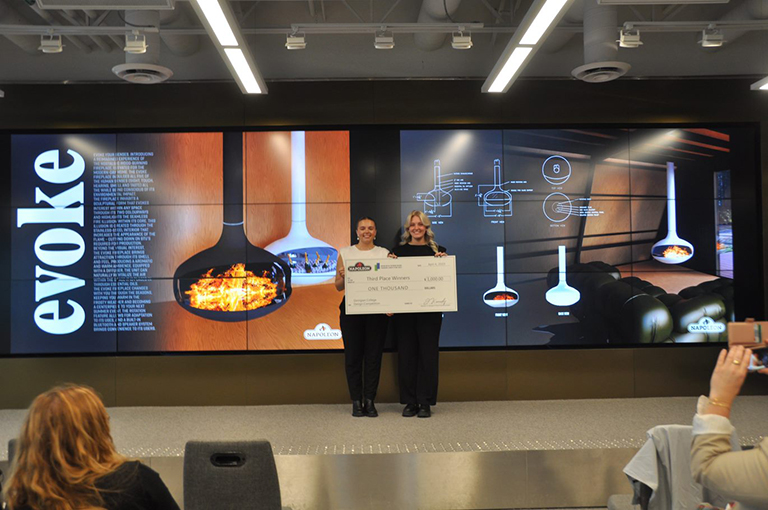 Two females stanging on a stage in front of a video screen. They're holding up a large cheque.