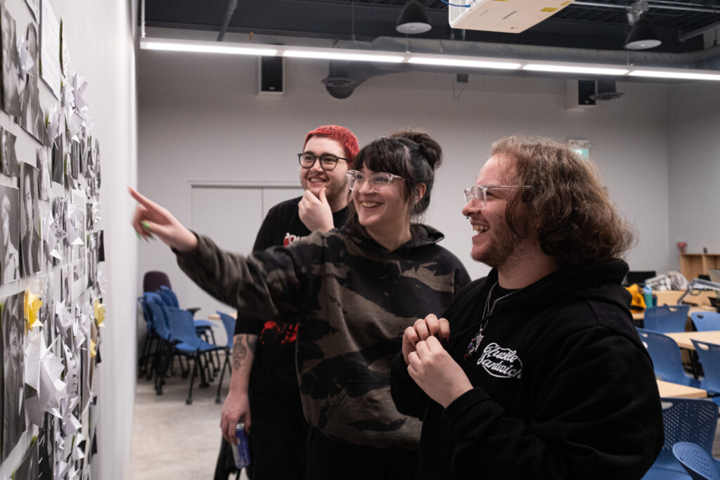 Three people stand in an art classroom and point at photos on a wall. 