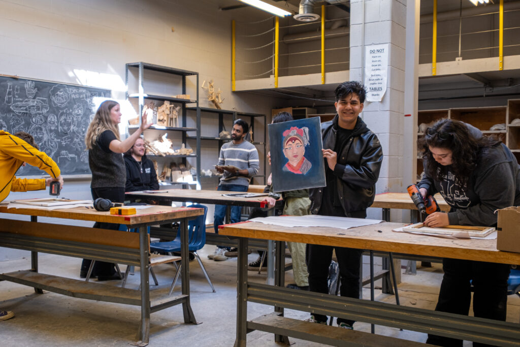 Several people stand in a workshop and use power tools to put together artwork frames. 