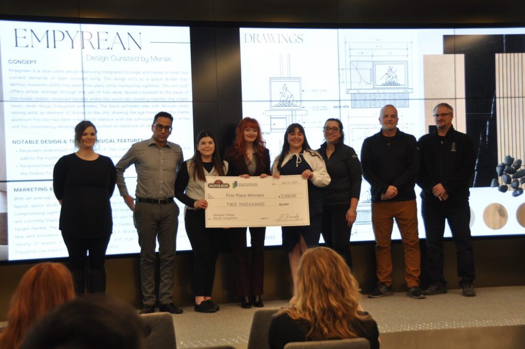 A group of eight people standing on a stage in front of a video screen. Three people in the centre are holding up a big cheque