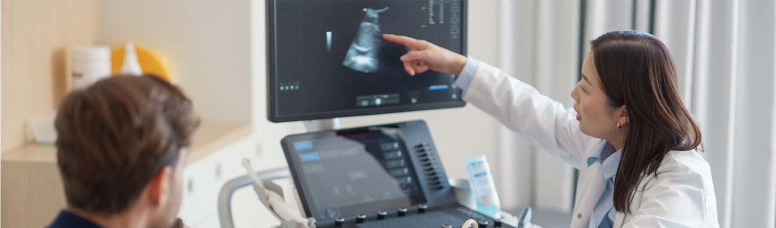 A medical professional wearing a white coat points at an ultrasound image on a monitor.
