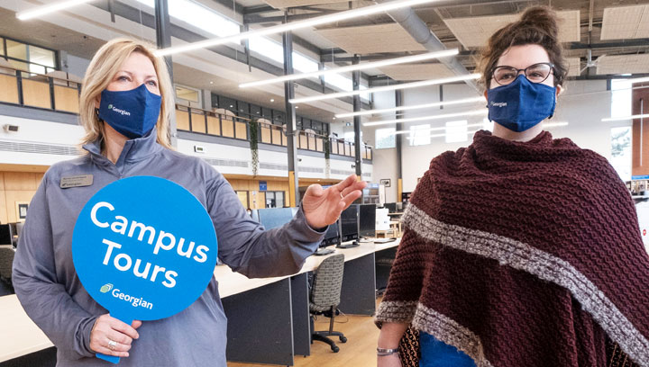 A Georgian student recruitment specialist holds a blue "Campus Tours" sign next to a student in the library at Georgian's Barrie Campus