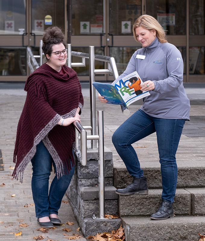 Sylvia Lauterbach, Student Recruitment Specialist at Georgian College, standing on steps outside the Barrie Campus main entrance while holding out an open viewbook during an interaction with a college student