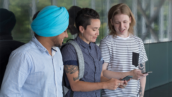 Three students leaning against a wall in M building at the Georgian College Barrie Campus smiling and looking at their cellphones