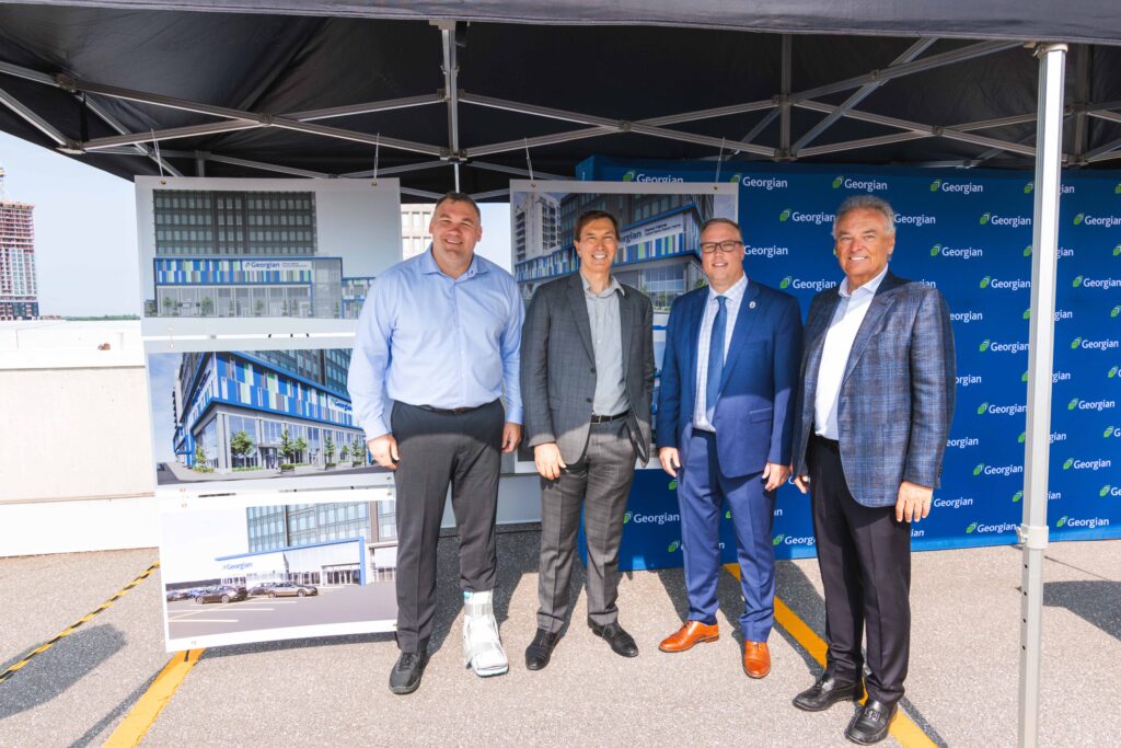 Four people wearing suits stand outside under a tent and next to a backdrop reading "СŶƵ" and several image boards of a building design.
