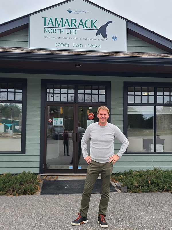 Person in a gray long-sleeve shirt and green pants in front of a building front entrace