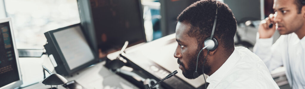 An emergency dispatcher with short, dark hair, wearing a headset and white long-sleeved collared shirt, using a phone system and switchboard to take an emergency phone calll