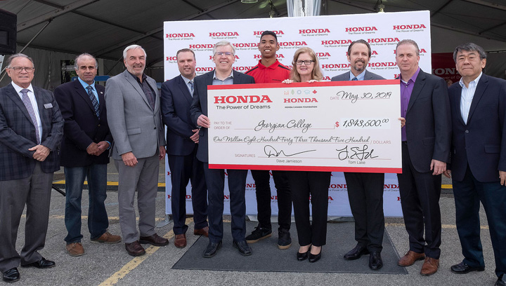 A group of men and women in business attire standing in front of a tent opening. They're holding up a large cheque from Honda in the amount of $1,843,500.