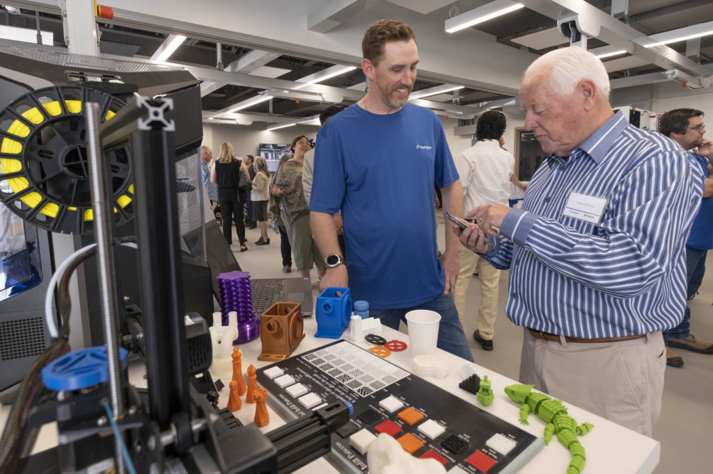 A white male wearing a blue t-shirt talking to an older white male. They're beside some electronic equipment.