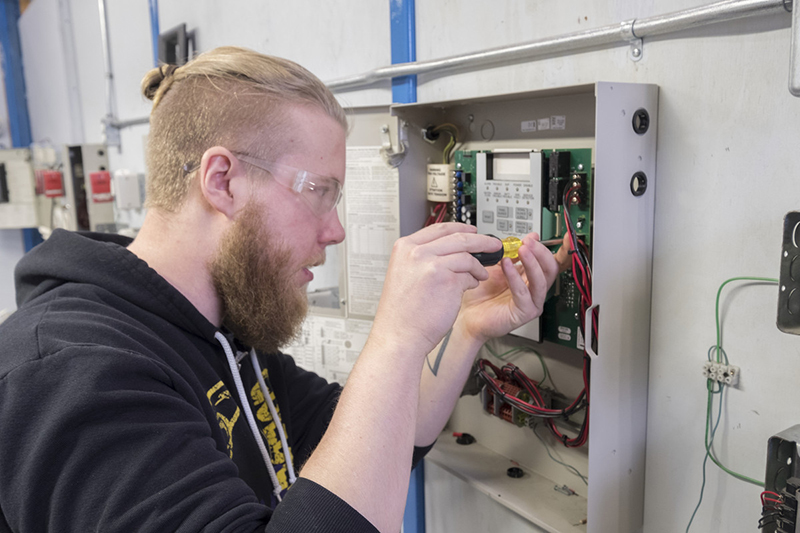 A male with short blonde hair and a beard wearing safety glasses. He's working on an electrical panel