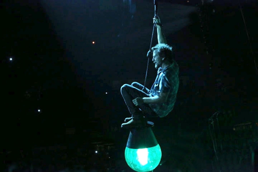 A person sits on top of a large blue light hanging from the ceiling of a concert venue. 