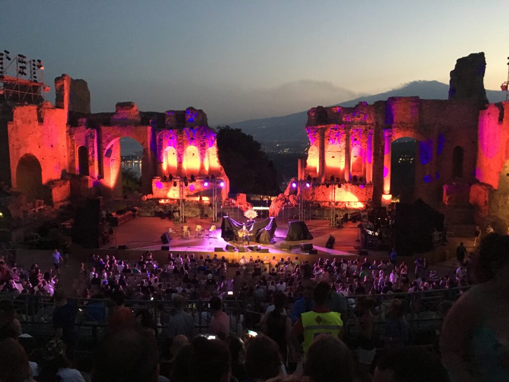 An outdoor venue with ancient-looking stone columns lit up in pink and yellow lights, with a band playing on stage and a mountain in the distance.