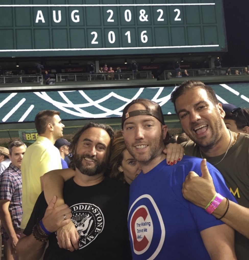 Four people put their arms around each other while standing in a crowd inside a baseball stadium. 