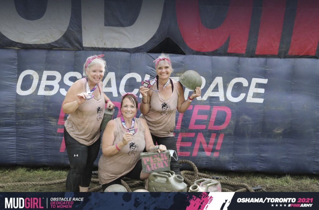 Three people pose together in front of a large sign. The image reads "Mud Girl Obstacle Race."