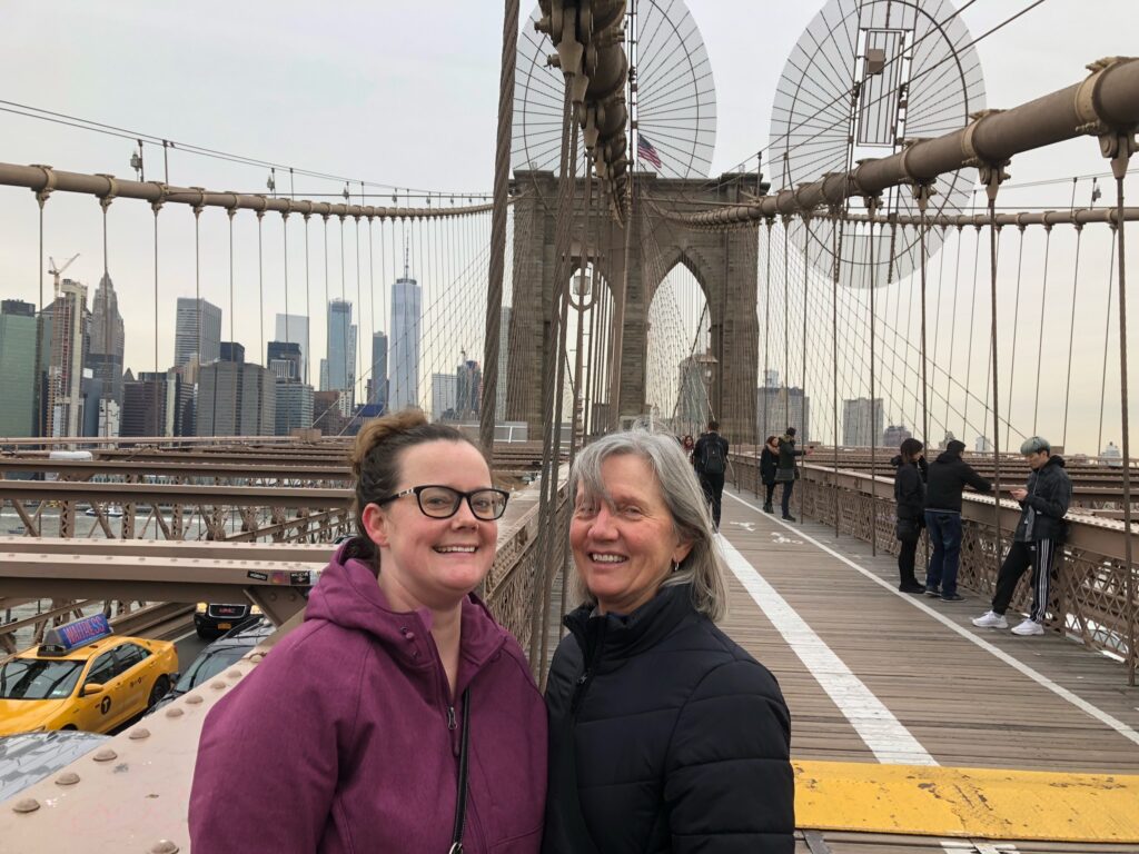 Two adults stand together and smile on the Brooklyn Bridge, with the New York skyline in the background. 