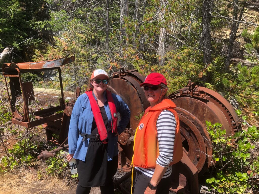 Two people walk through a wooded area near abandoned, rusted equipment.